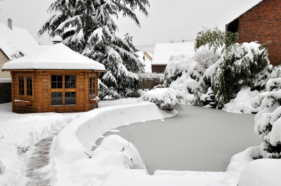 Verschneites Gartenhaus neben zugefrorenem Teich